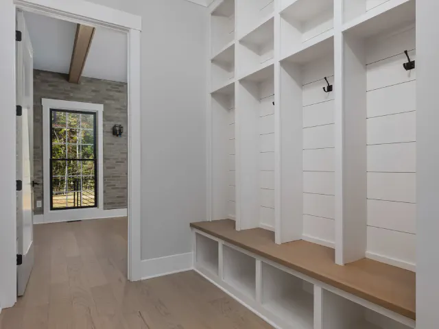 Biała zabudowa typu mudroom z otwartymi schowkami, haczykami, drewnianym siedziskiem i przegródkami na buty pod spodem