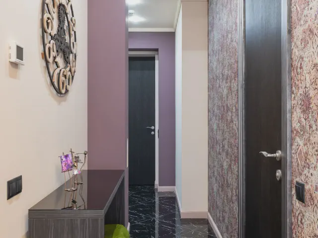 Korytarz z jedną ścianą w przygaszonym, liliowo-różowym kolorze, czarnymi drzwiami, zegarem ściennym, ciemną konsolą i wzorzystą tapetą obok