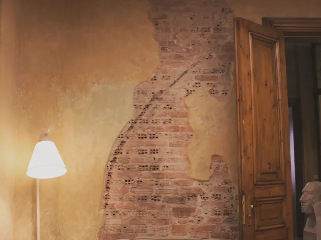 Fragment ściany z odsłoniętą czerwoną cegłą spod starego tynku, obok drewniane drzwi z płycinami, jasny fotel i lampa podłogowa