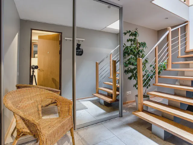 Hol w domu z drewnianymi schodami na metalowej balustradzie, szafą z lustrzanymi drzwiami, rattanowym fotelem i dużą rośliną w doniczce
