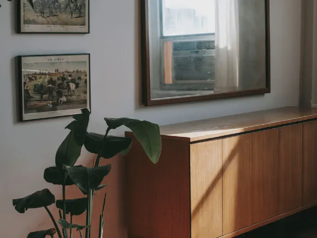 Niska drewniana komoda w stylu mid-century pod lustrem w ciemnej ramie, obok stara fotografia w ramce i duża roślina