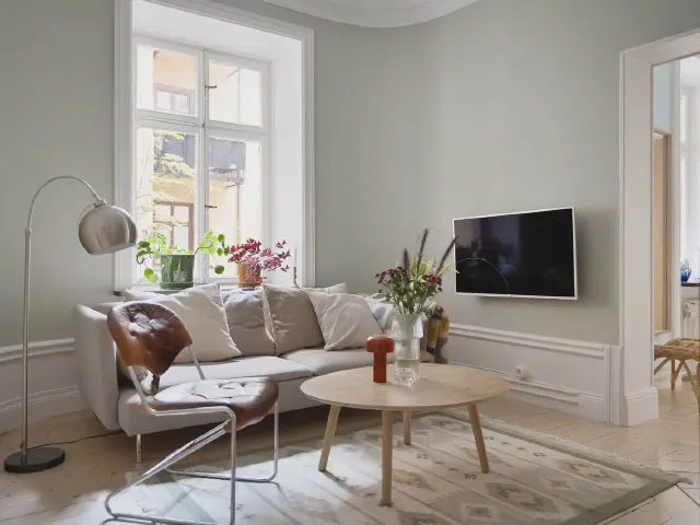 Jasny mały salon z szarą sofą, okrągłym drewnianym stolikiem, lampą łukową i telewizorem na ścianie