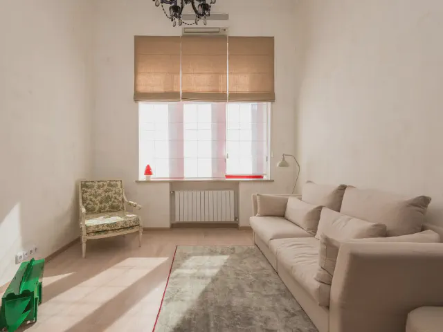 Mały salon z beżową sofą, kwiecistym fotelem przy oknie z rzymską roletą i miękkim dywanem