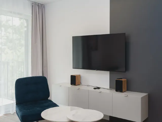 Telewizor na grafitowym fragmencie ściany nad białą wiszącą szafką z głośnikami, obok granatowy fotel