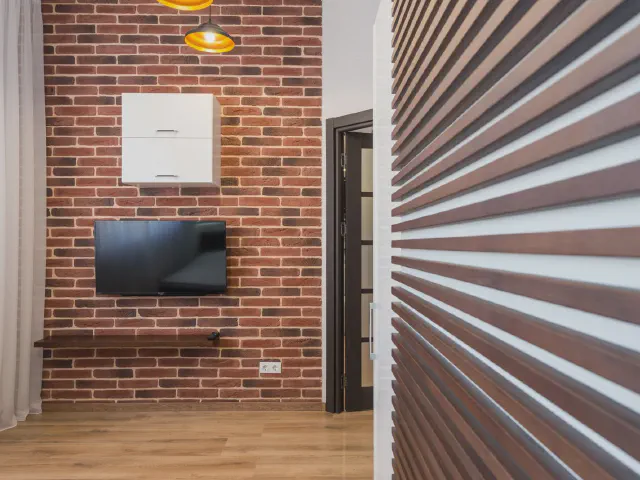 Telewizor na ścianie z czerwonej cegły z drewnianą półką, na pierwszym planie ściana z drewnianych listew
