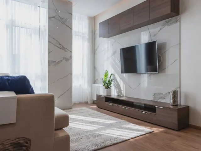 Telewizor na ścianie z wielkoformatowych płyt imitujących biały marmur, pod nim długa szafka RTV z orzecha i wiszące szafki