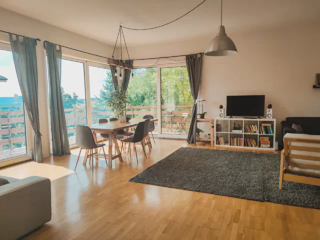 Jasny salon z jadalnią przy narożnych przeszkleniach, szare zasłony, drewniany stół i wyjście na długi balkon