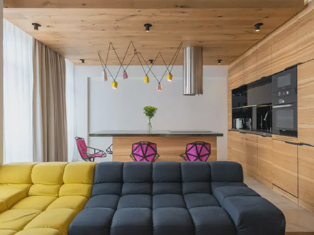 Kuchnia z wyspą i zabudową z jasnego drewna na całą ścianę, przed nią żółto-granatowa modułowa sofa, nad wyspą kolorowe lampy