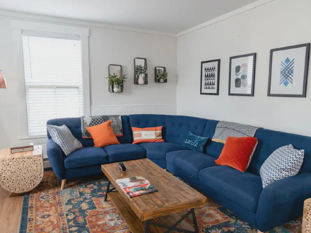 Granatowy narożnik z pomarańczowymi i wzorzystymi poduszkami, drewniany stolik kawowy i kolorowy dywan