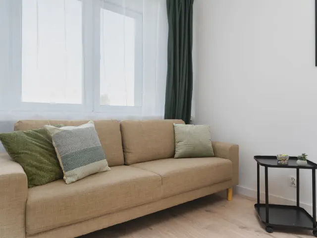 Beżowa sofa trzyosobowa z poduszkami w butelkowej zieleni i szarości, obok czarny metalowy stolik pod oknem z zieloną zasłoną