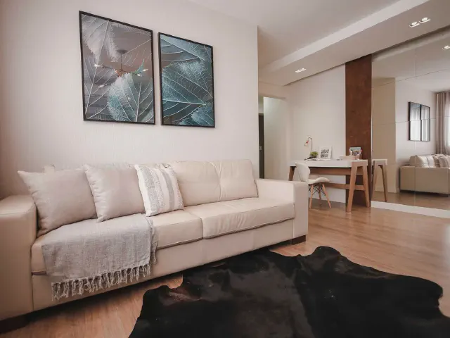 Jasna beżowa sofa z kremowym pledem i poduszkami, dwie grafiki z motywem liści nad kanapą, czarna skóra zwierzęca na drewnianej podłodze