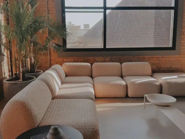 Modułowa beżowa sofa z niskich segmentów przed ścianą z czerwonej cegły, duże okno w czarnej ramie i palmy w donicach