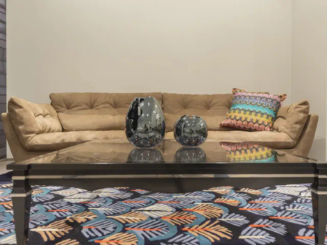 Beżowa pikowana sofa z kolorową poduszką w geometryczny wzór, szklany stolik z ciemnymi wazonami i wielobarwny dywan w liście