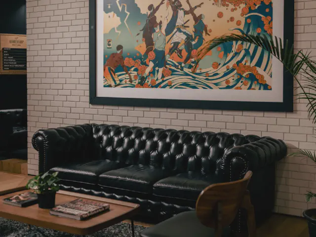 Czarna pikowana sofa Chesterfield przed ścianą z białej cegły, nad nią duży kolorowy obraz, na pierwszym planie drewniany stolik