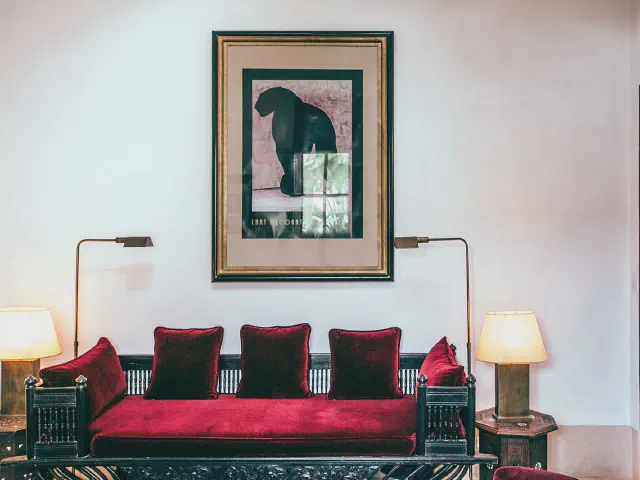 Czerwona welurowa sofa w stylu kolonialnym z ciemną rzeźbioną ramą i bordowymi poduszkami, po bokach lampy z abażurami, nad nią oprawiona grafika