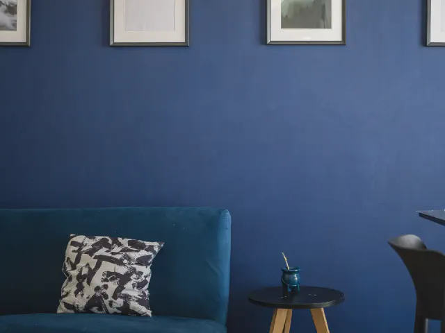 Ciemnoniebieska welurowa sofa z biało-czarną poduszką na tle ściany w głębokim granacie, nad nią grafiki w czarnych ramach