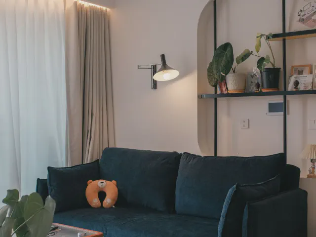 Granatowa sofa z pomarańczową poduszką w salonie z ciepłymi beżowymi ścianami, półkami z roślinami i jasnymi zasłonami