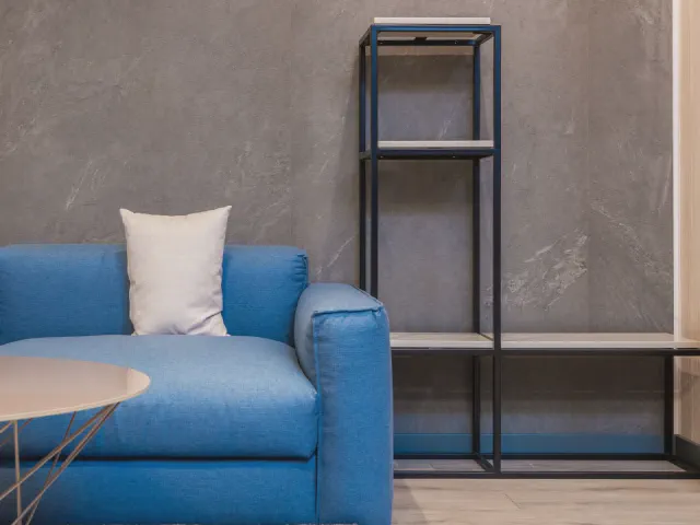 Intensywnie niebieska sofa z białą poduszką przy szarej ścianie w dekorze betonu i czarnym metalowym regale
