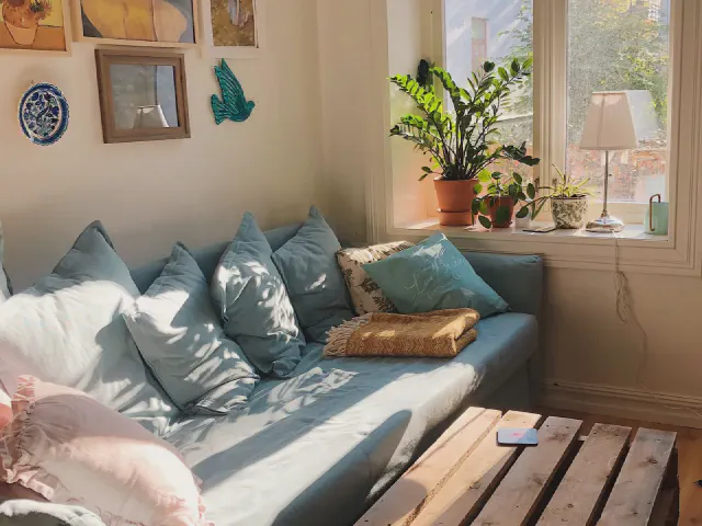 Błękitna sofa z wieloma poduszkami w odcieniach niebieskiego i różu, stolik z palet, rośliny w donicach na parapecie i ceramiczne dekoracje na ścianie