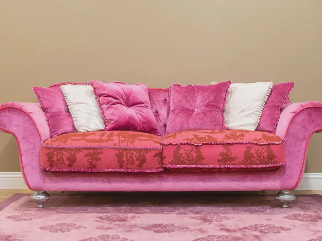Klasyczna sofa w intensywnym różu z welurowym obiciem, poduszkami w różu i bieli oraz siedziskami w czerwony wzór, na różowym dywanie przy beżowej ścianie