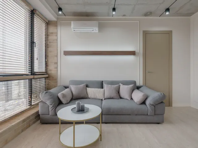 Popielata sofa z pluszowymi poduszkami w jasnym salonie z żaluzjami na przeszklonej ścianie, okrągłym białym stolikiem na złotej nodze i betonowym sufitem