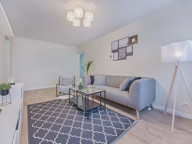 Szara sofa na drewnianych nóżkach, granatowy dywan w marokańską koniczynę, metalowy stolik kawowy i lampa podłogowa z trójnogiem