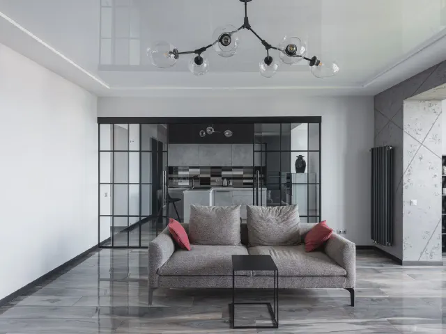 Szara sofa o prostej formie z czerwonymi poduszkami w dużym salonie z połyskującą podłogą, szklaną ścianą w czarnych ramach i nowoczesnym żyrandolem