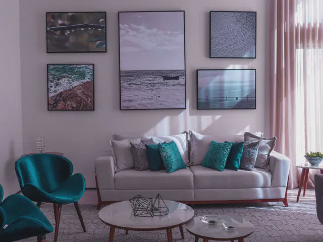 Biała sofa z turkusowymi i szarymi poduszkami, turkusowe fotele, okrągłe stoliki i fotografie morza na jasnej ścianie