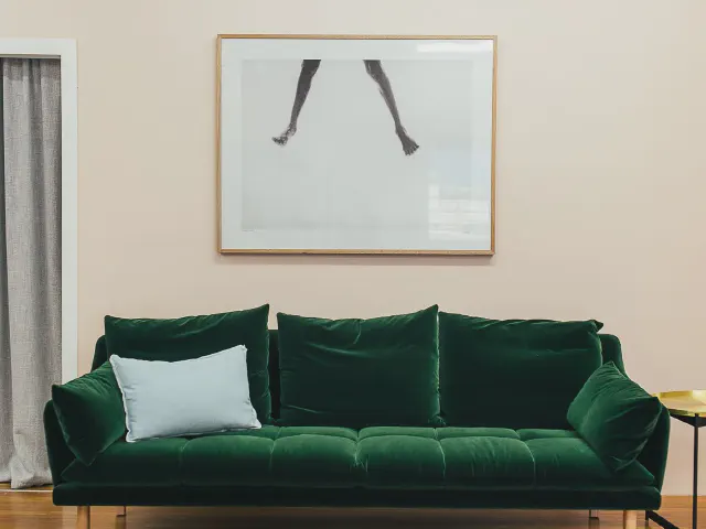 Welurowa sofa w butelkowej zieleni z jasnoniebieską poduszką, nad nią grafika w jasnej ramie na tle ciepłej, jasnobeżowej ściany
