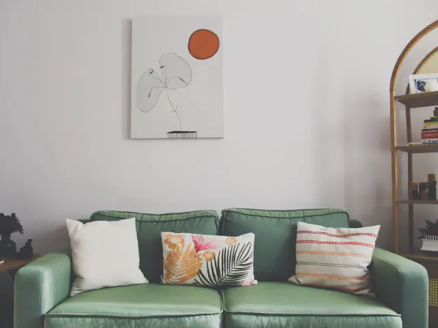 Sofa w szałwiowej zieleni z białą i wzorzystymi poduszkami, nad nią grafika z liściem, obok regał w łukowej ramie
