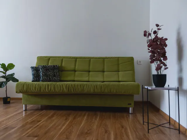 Oliwkowa sofa z pikowanym siedziskiem i ciemnymi poduszkami w jasnym pokoju z drewnianą podłogą, metalowym stolikiem i roślinami w donicach