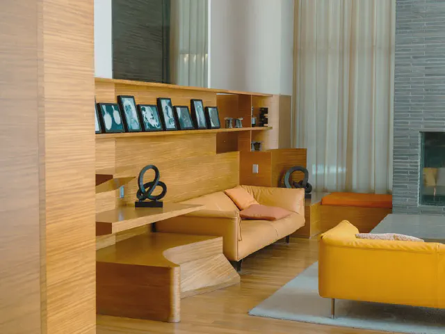 Salon z drewnianą zabudową i półkami, bursztynowa skórzana sofa, intensywnie żółty fotel, szara ściana z kominkiem i wysokie zasłony