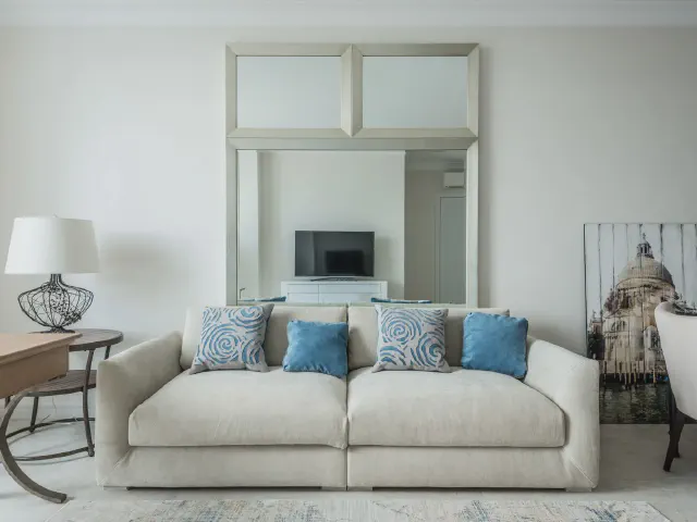 Jasna sofa w odcieniu ecru z niebieskimi i wzorzystymi poduszkami na tle białej ściany z lustrem