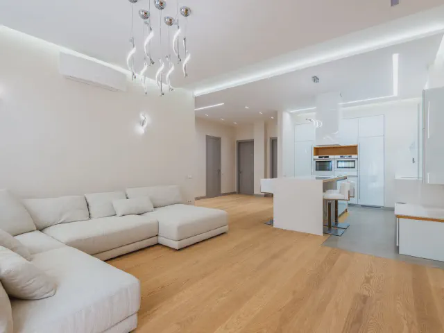 Otwarty salon z białymi ścianami, dużym białym narożnikiem, dębową podłogą i białą kuchnią z wyspą w tle
