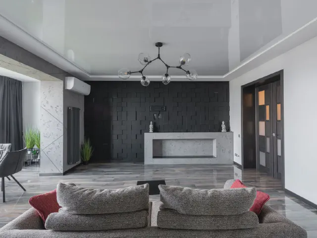 Salon z czarną ścianą z paneli 3D, jasną zabudową kominkową, szarymi sofami z czerwonymi poduszkami i białym sufitem