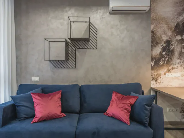 Granatowa sofa z czerwonymi, satynowymi poduszkami na tle szarej ściany z metalową półką