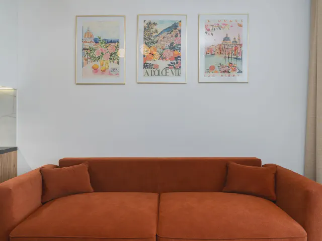 Sofa w odcieniu rdzawej, ceglanej czerwieni pod białą ścianą z trzema kolorowymi plakatami