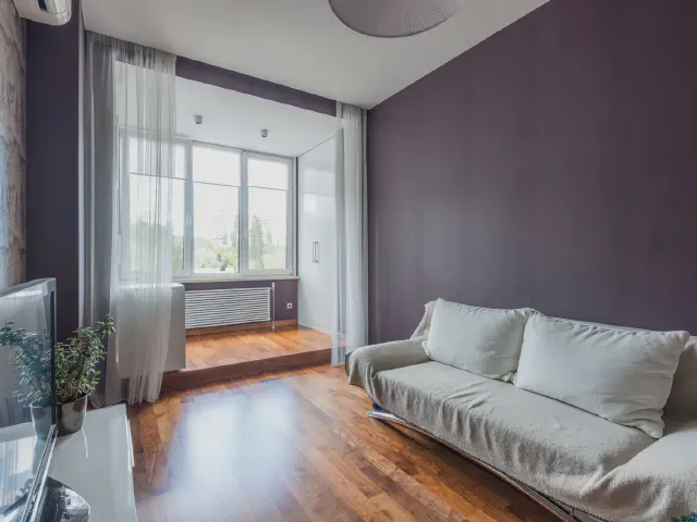 Salon ze ścianą w głębokim, bakłażanowym fiolecie, jasnoszarą sofą, drewnianą podłogą i dużym oknem z białymi firanami