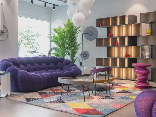Nowoczesny salon z fioletową pikowaną sofą o obłych kształtach, kolorowym dywanem w geometryczne wzory i podświetlanym regałem