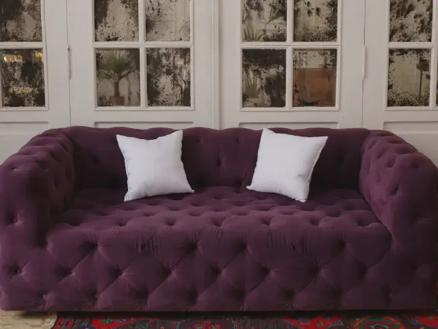Śliwkowa, welurowa sofa pikowana w stylu chesterfield z dwiema białymi poduszkami