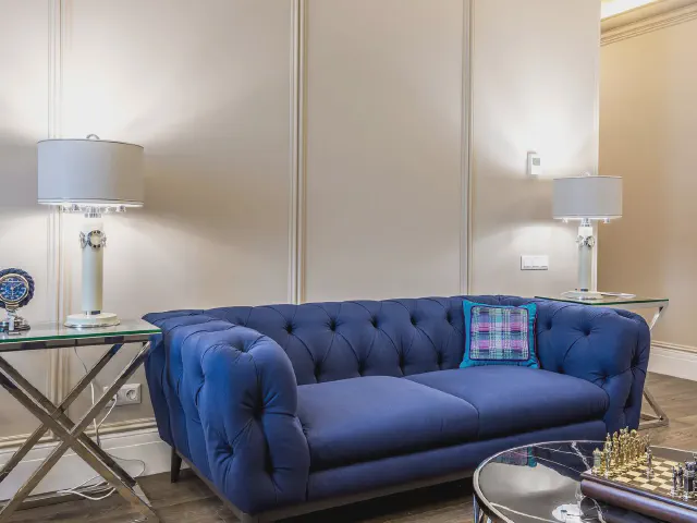 Pikowana sofa chesterfield w nasyconym, szafirowym odcieniu granatu na tle kremowych ścian z listwami, szklane stoliki i białe lampy