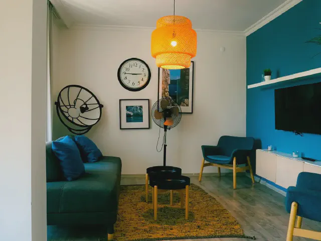 Salon z ciemnoturkusowo-niebieską ścianą z telewizorem, niebieską sofą i fotelami, żółtym dywanem i pomarańczową lampą wiszącą