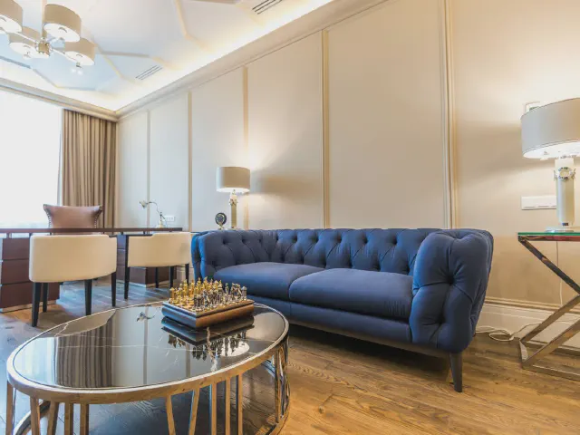 Granatowa pikowana sofa w jasnym salonie z beżowymi ścianami, szklanym stolikiem kawowym i złotymi detalami