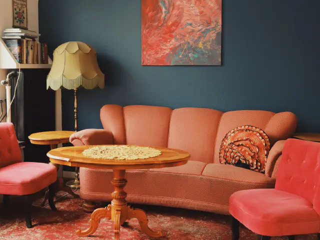 Łososioworóżowa welurowa sofa, czerwone fotele, drewniany okrągły stolik i lampa z abażurem na tle ciemnej, zielononiebieskiej ściany