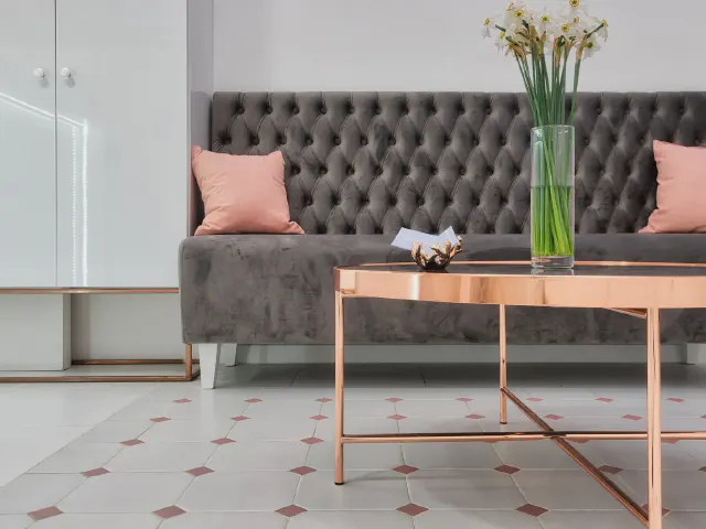 Szara pikowana sofa z różowymi poduszkami, stolik kawowy w kolorze różowego złota z wazonem żonkili i posadzka w romby