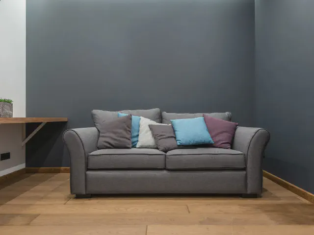 Szara sofa z błękitnymi, białymi i śliwkowymi poduszkami przed grafitową ścianą, drewniana podłoga i półka-biurko przy ścianie