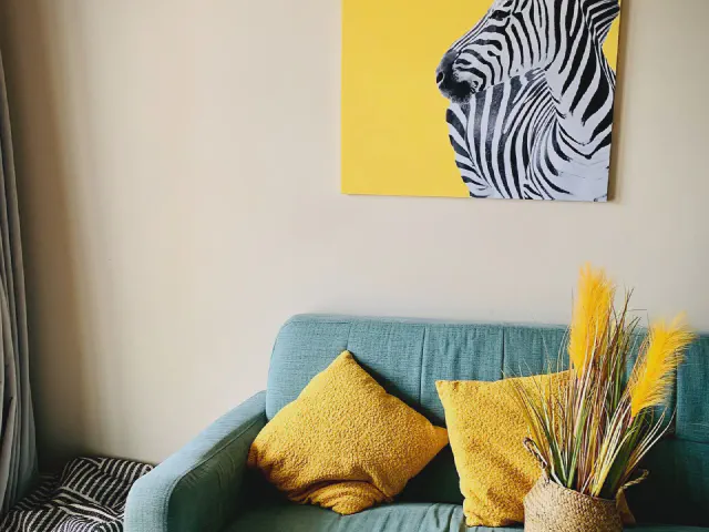 Turkusowa sofa z musztardowymi poduszkami, suszone trawy i obraz z zebrą na żółtym tle przy kremowej ścianie