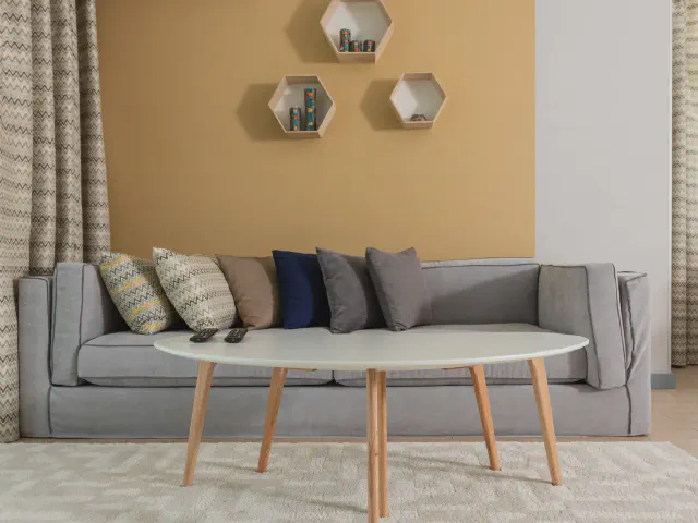 Szara sofa z wzorzystymi i granatowymi poduszkami przed ścianą w kolorze ochry z heksagonalnymi półkami, owalny stolik na drewnianych nogach