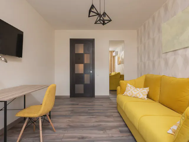 Żółta kanapa i żółte krzesło przy biurku w jasnym salonie z szarą podłogą i lampą z geometrycznym kloszem