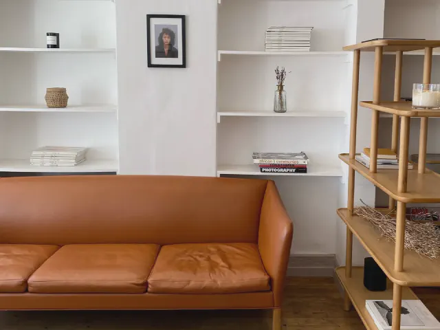 Brązowa skórzana sofa przed wnęką z białymi półkami, obok drewniany regał z otwartymi półkami